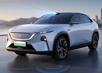 Front three-quarter view of a white Mazda EZ-60 LiDAR variant with a roof-mounted LiDAR sensor emitting blue light beams, parked on a waterfront road with mountains and a city skyline in the background. Blue rings on the ground illustrate the vehicle's sensor coverage.
