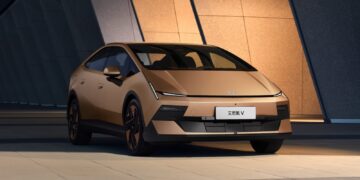 Gold Hyundai Ioniq V electric sedan viewed from the front three-quarter angle outside a modern building.
